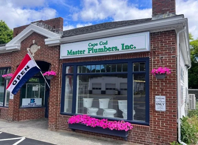 The brick Cape Cod Master Plumbers storefront in Sagamore with pink flower boxes and toilets on display in the window